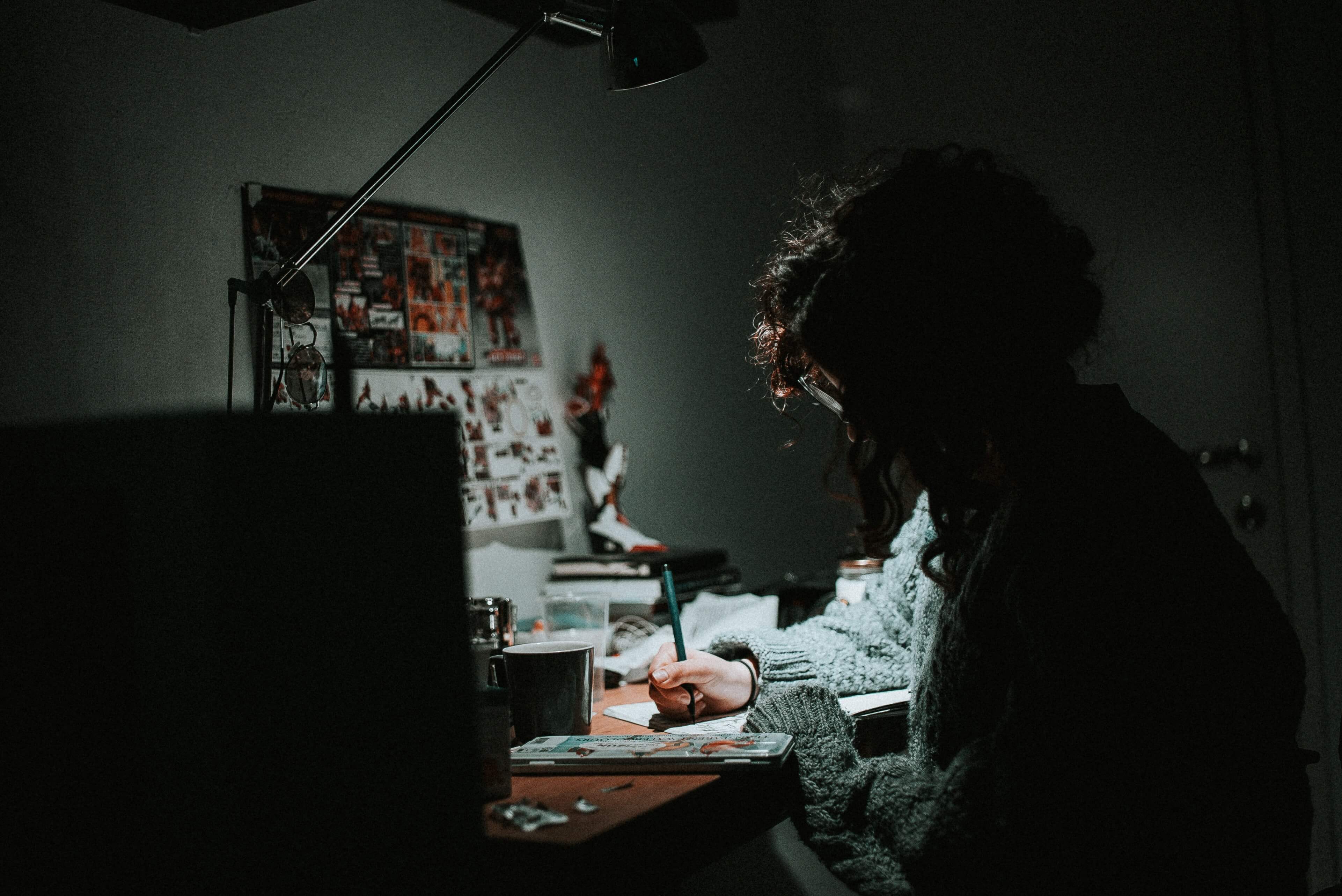 Female drawing at a desk in a dimly lit room with a mood board above and a green mug on the desk.