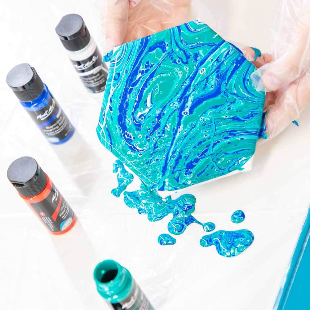 3. Hands holding a hexagonal canvas with blue pour paints on it and SuperCell paints in the background.