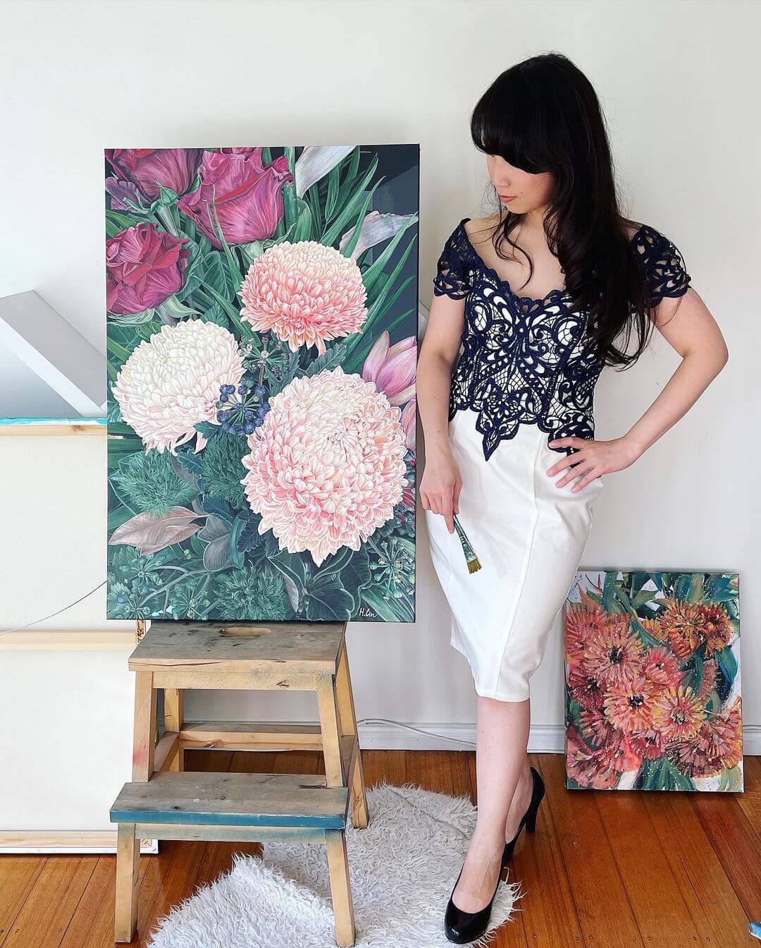 Hsin Lin standing happily next to her canvas holding a paint brush in her right hand with a realistic painting of flowers.