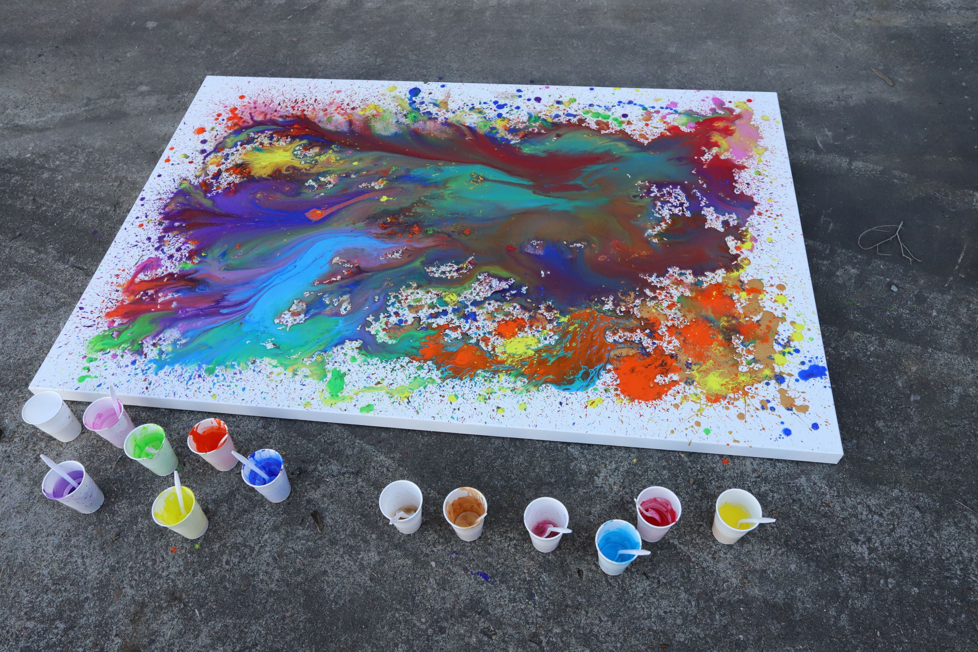 3. Rainbow coloured drip painting on a large white canvas with paints in cups around