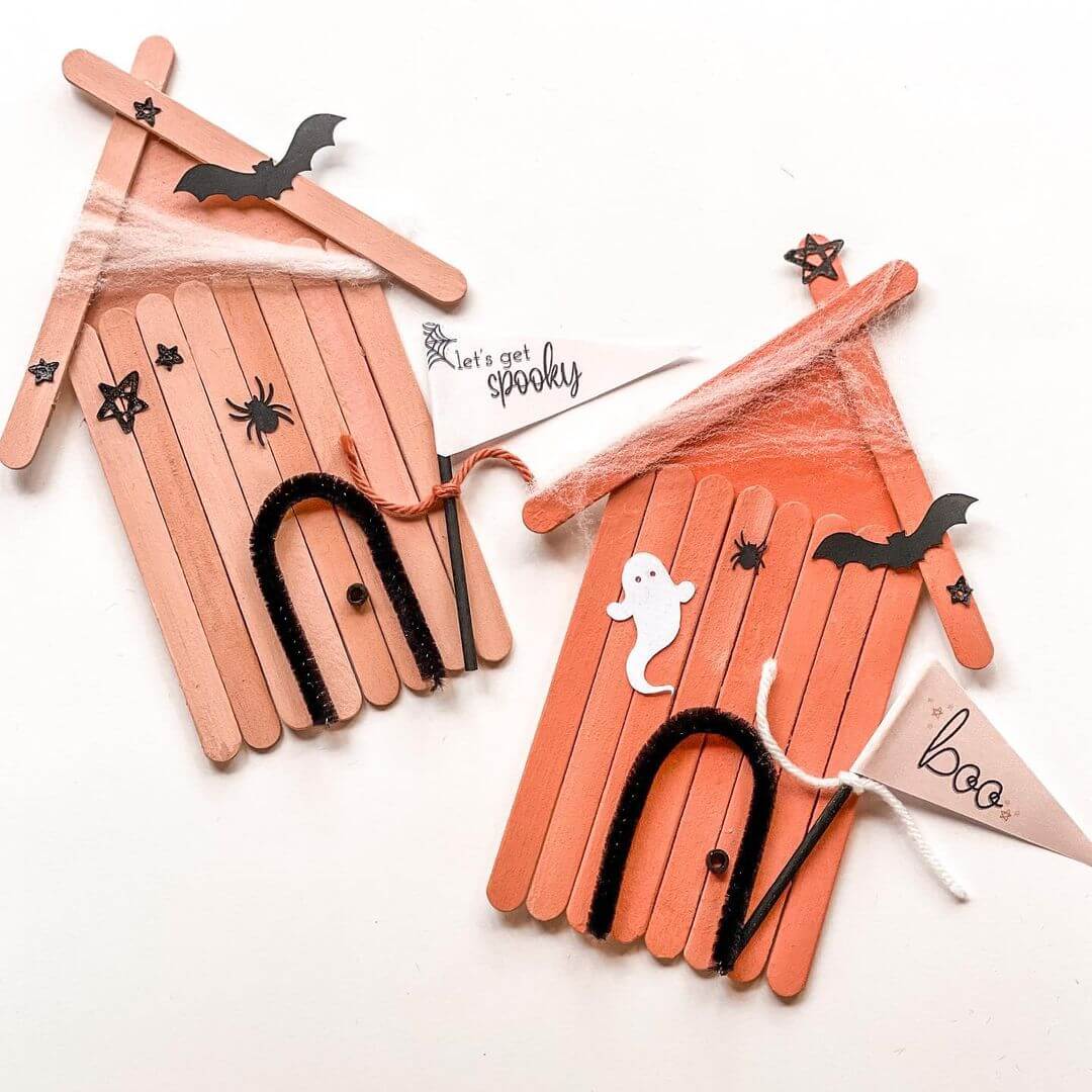 Two haunted houses made using popsicle sticks are stuck on a piece of white paper with spooky decal stickers and flags on the front of the house.