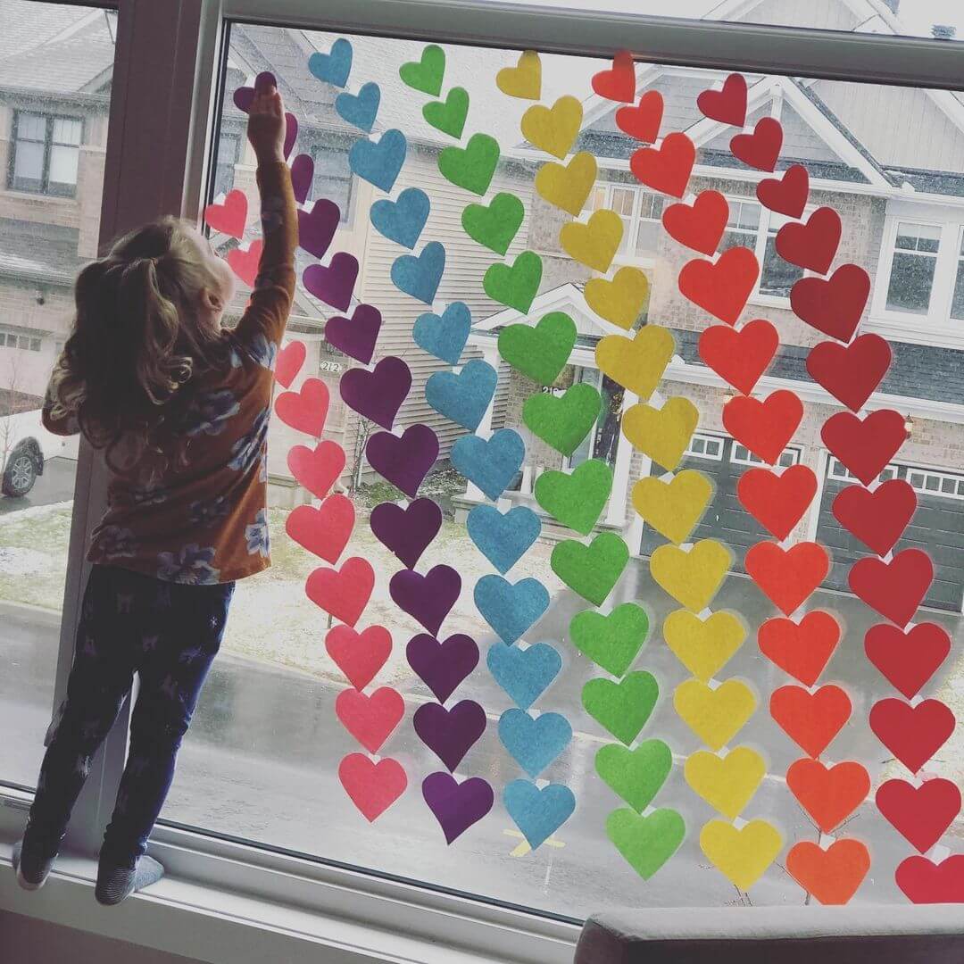 Young girl sticking a purple heart on a row of coloured rainbow hearts on a window.