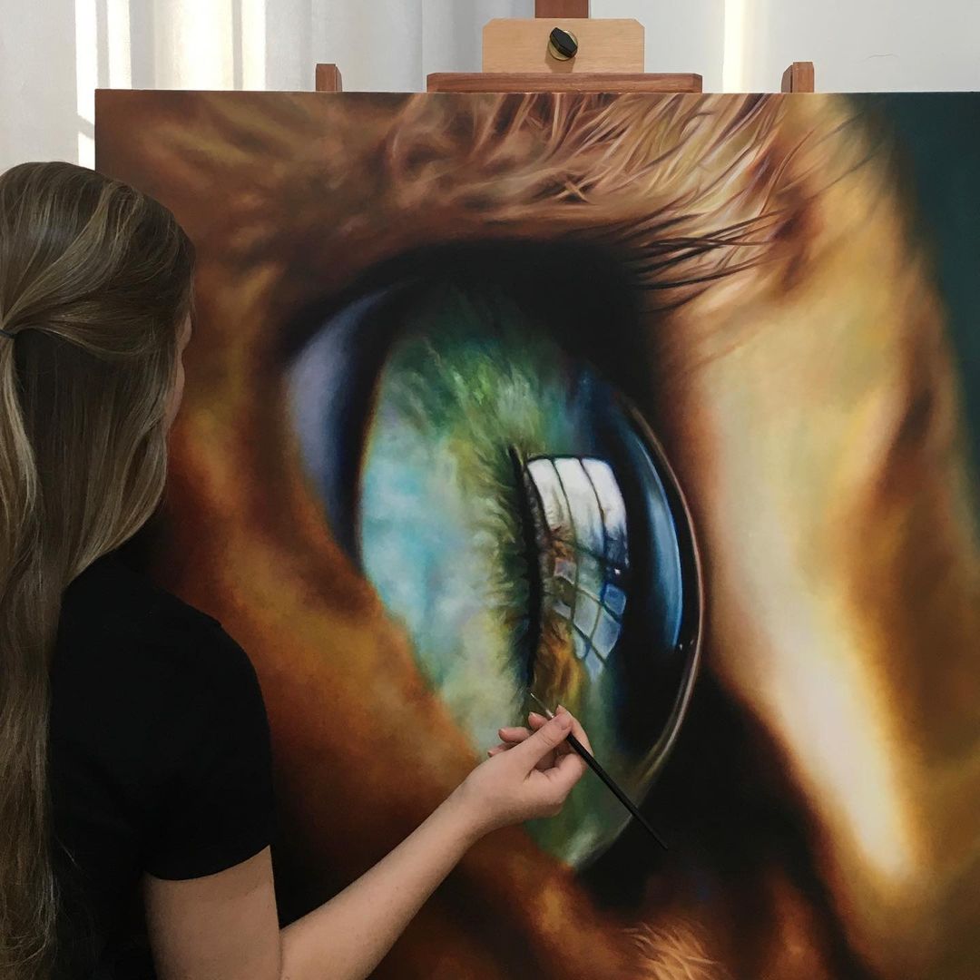 3. @ligiafiedler standing in front of her painting of an orange cat's green eye in hyperealistic detail