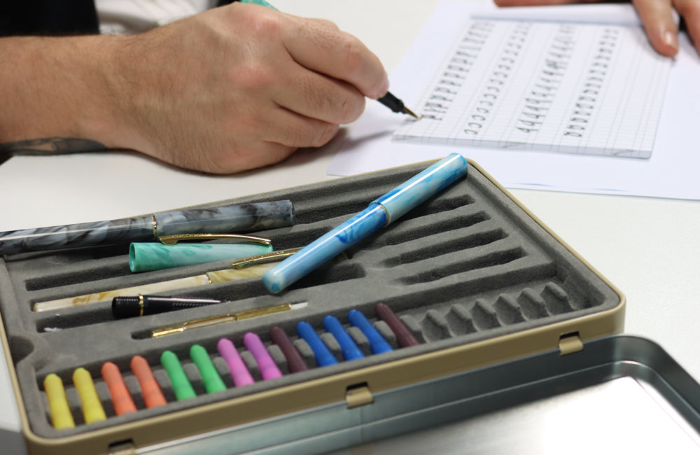 Case of fountain pens and ink cartridge's next to a hand writing calligraphy.