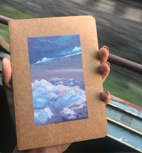 Hand holding a gouache artwork of clouds on kraft paper.