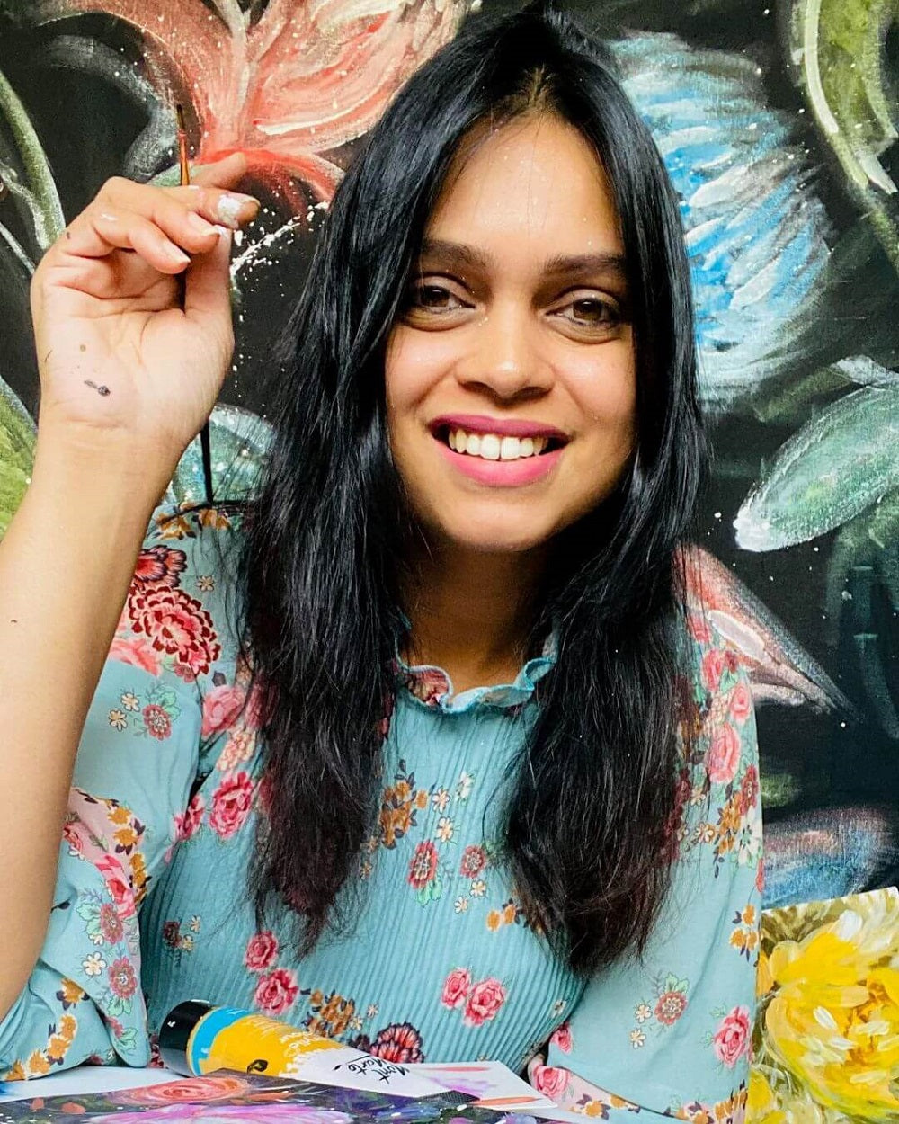 Artist Pallavi holding a paint brush and smiling in a blue and pink floral dress with flower art behind her.