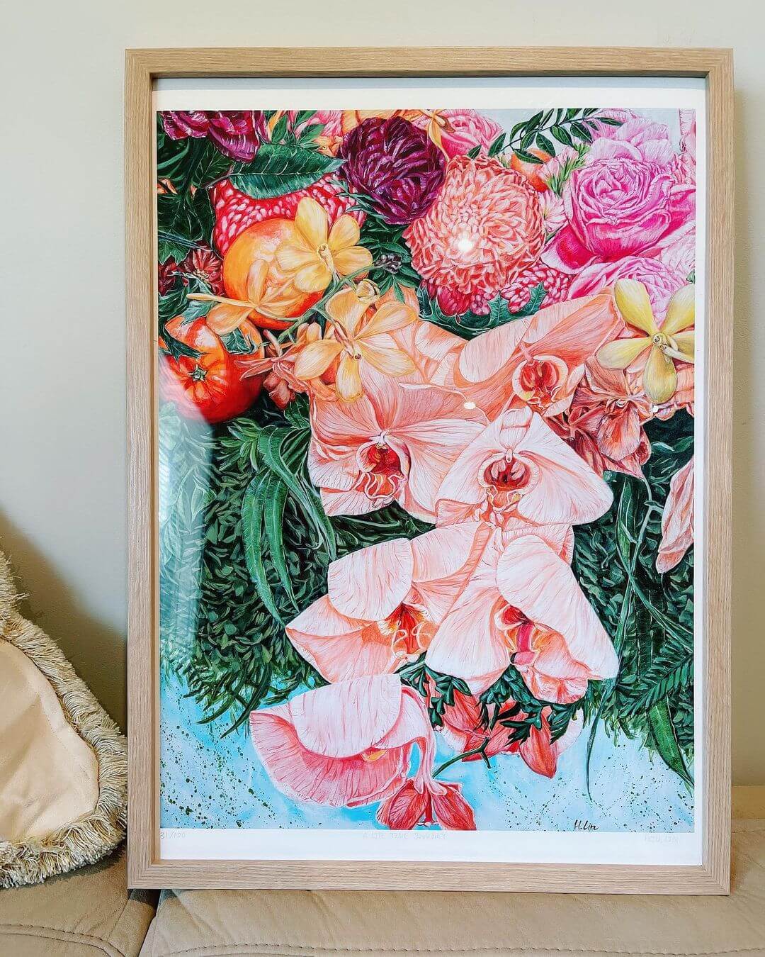 A framed artwork of colourful flowers painted in acrylic standing on a beige couch next to a pillow.