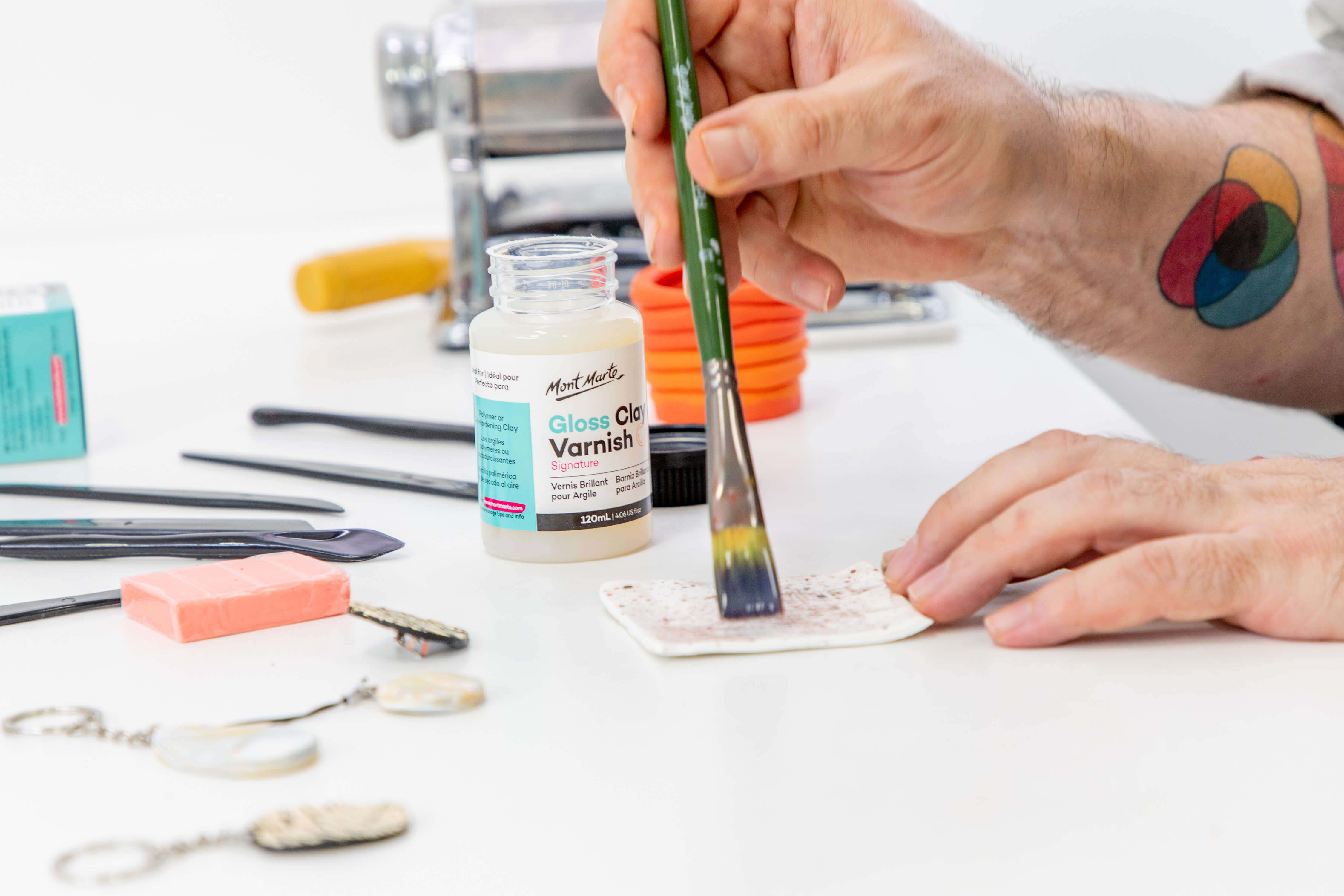 A hand uses clay varnish on a paintbrush to varnish polymer clay with various clay tools around.