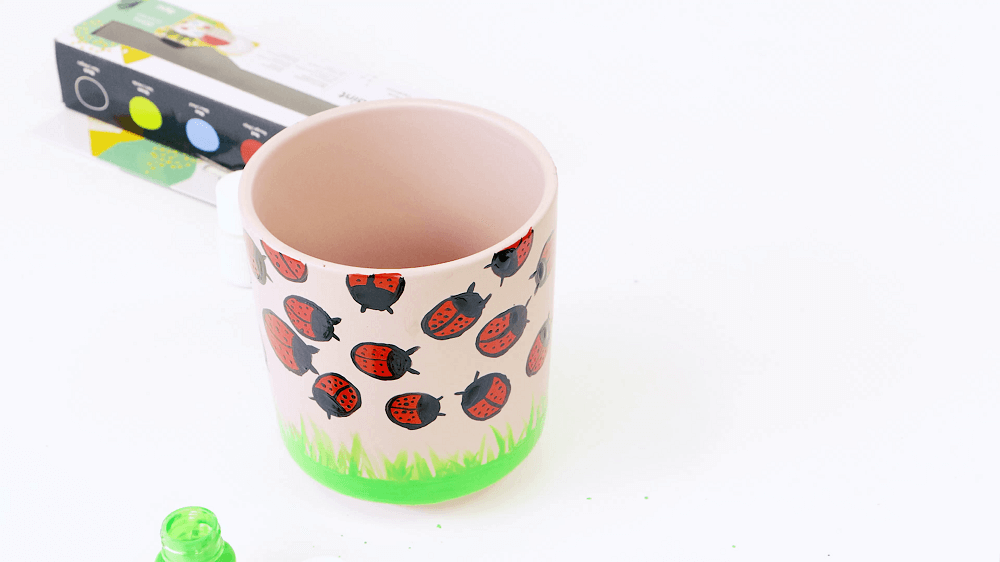 A pink ceramic glazed pot with red lady bugs painted on top and green grass.