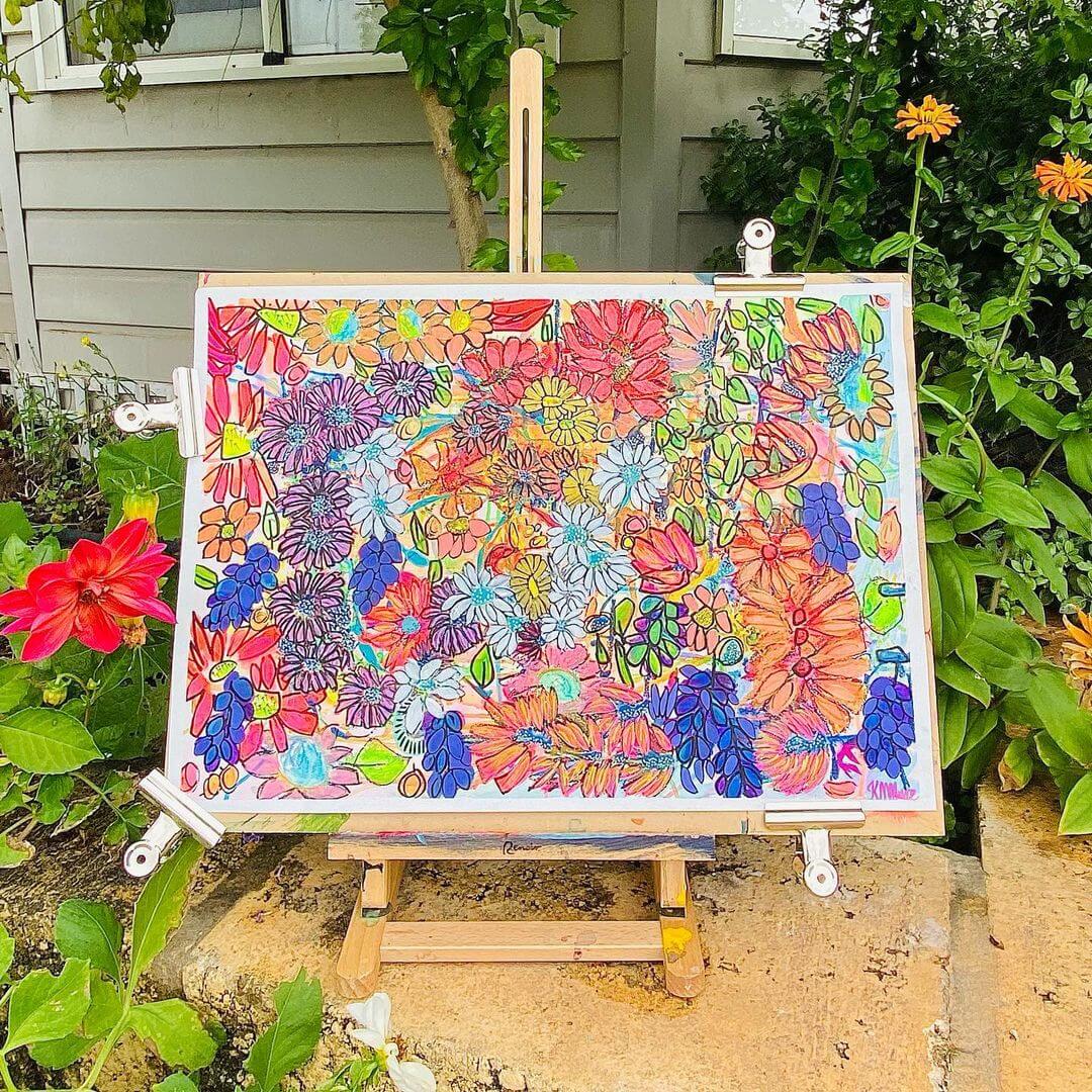 A whimsical, abstract artwork of flowers on paper. It's sitting on an easel with a farm house in the background.