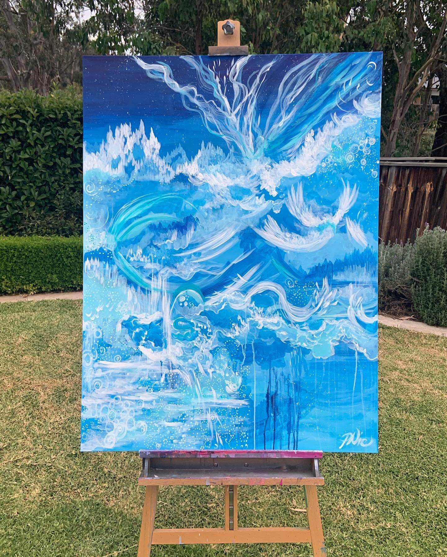 An easel with a canvas painting outside in a green backyard of Sydney. The canvas has blue paint painted in an abstract way with white waves.