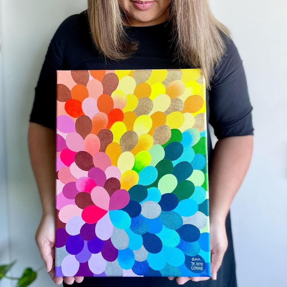 Ann in a black dress holding a colourful canvas with a petal design.