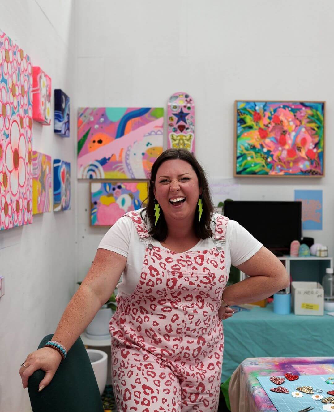 4. Artist Claire Monique laughing in her studio, wearing  pink leopard print overalls and yellow lightning bolt earrings.