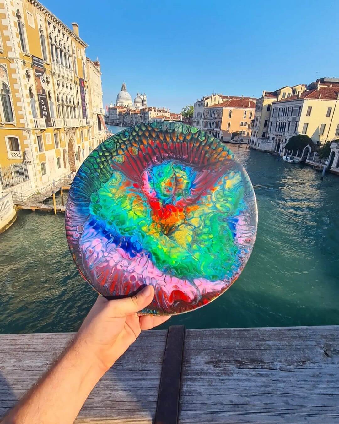 4. Hand holding a finished acrylic pour painting on a round circle, holding the work on a bridge in Venice.