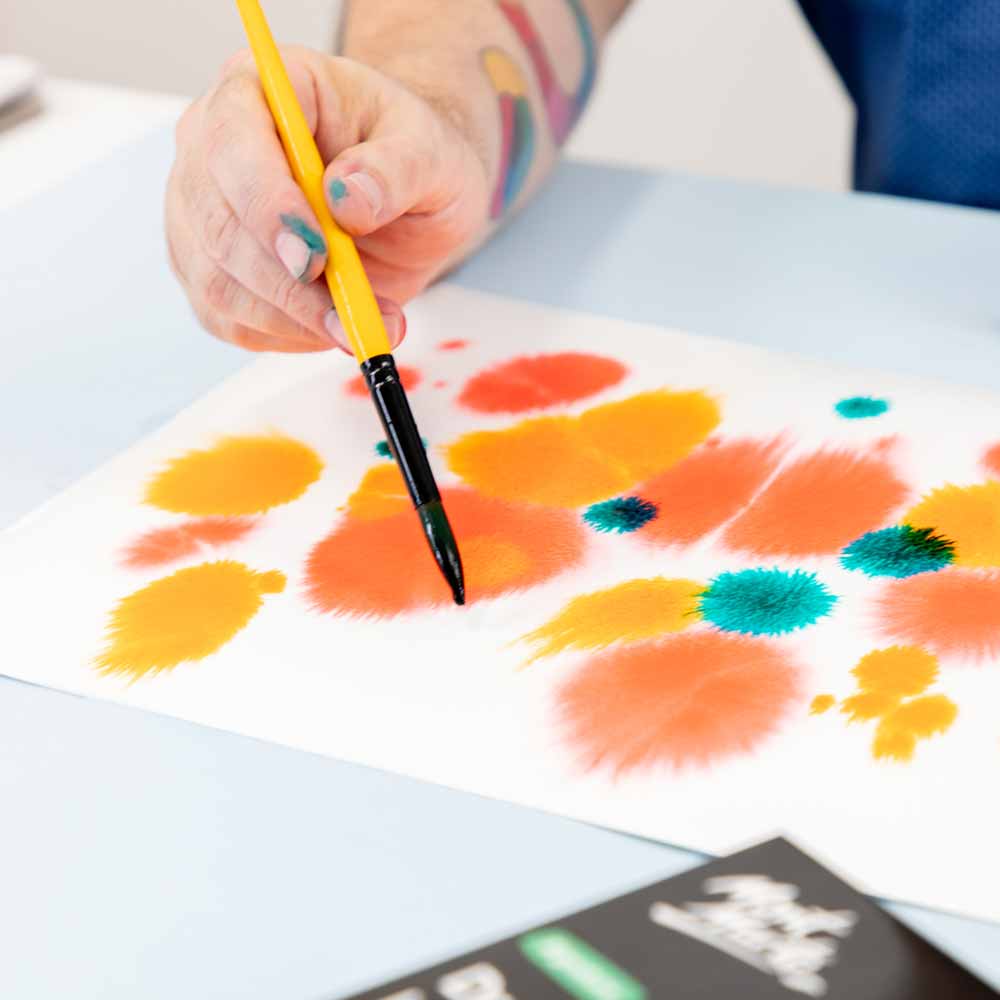 Hand holding a yellow brush and painting orange splats with drawing inks on white paper.