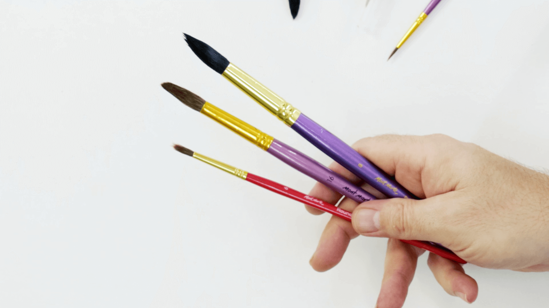 Hand holding three round watercolour brushes of different sizes.