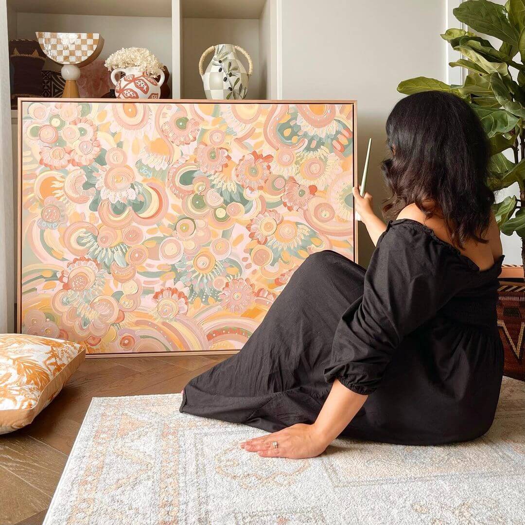 Lani sitting on a light coloured mat wearing a black dress and holding a completed artwork with pastel colours.