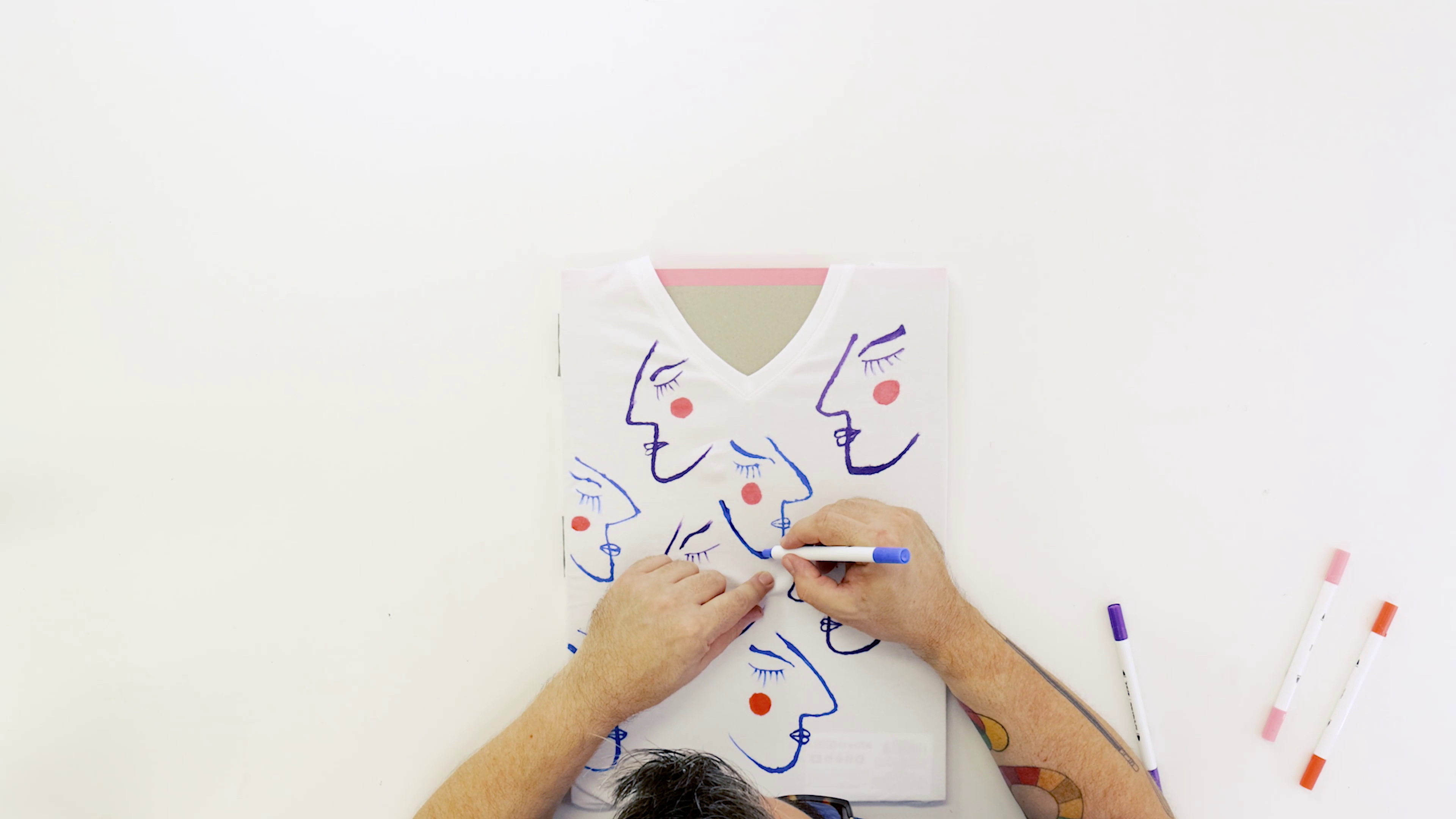 Man drawing abstract faces on a white tshirt with fabric paint markers.