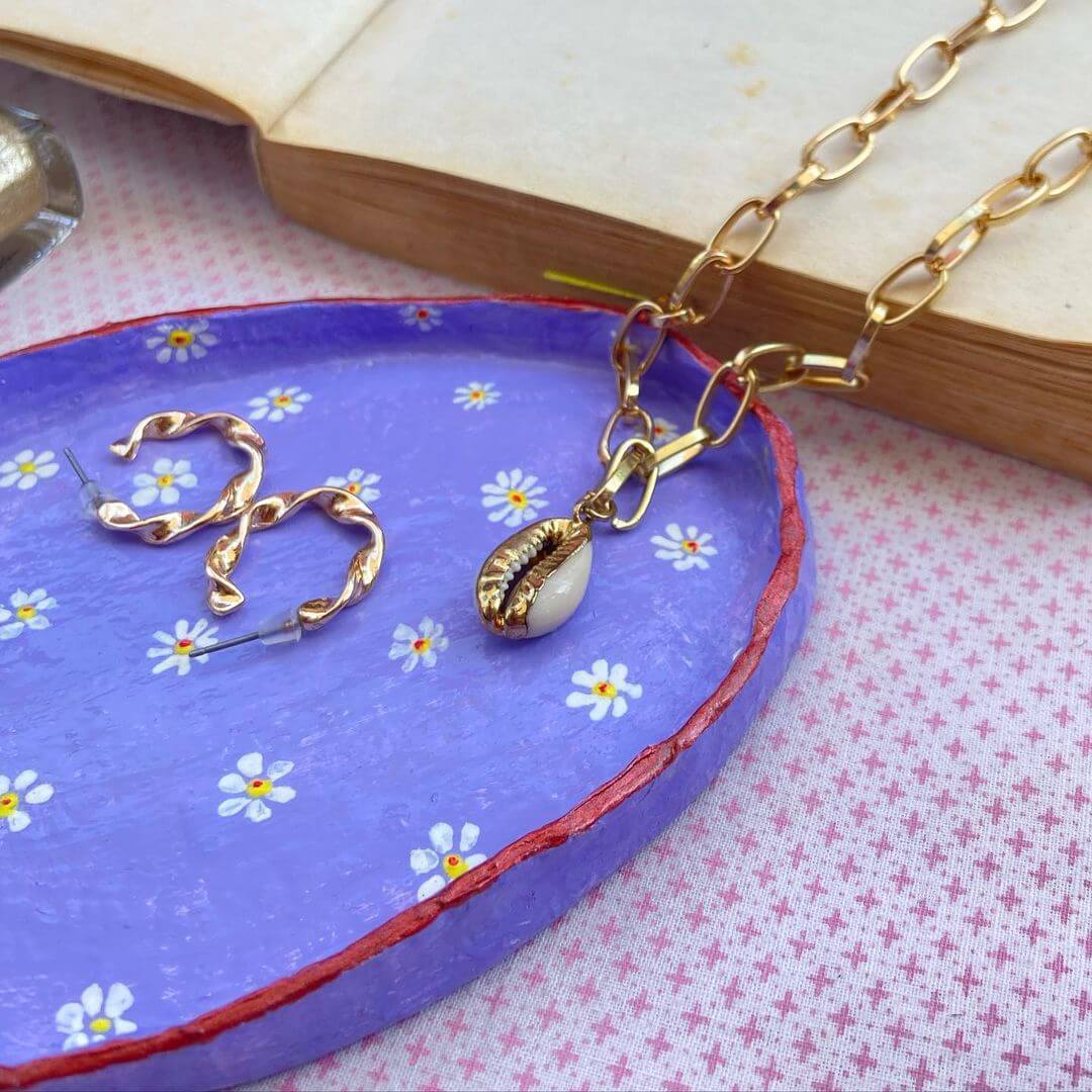 5. A clay trinket dish painted purple with white daisys painted on top. A shell necklace and gold hoop earrings sit on the tray.