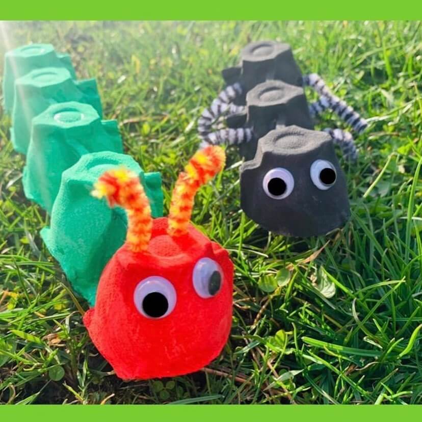 A red and green caterpillar made from an egg carton next to a black ant made from an egg carton on the grass.