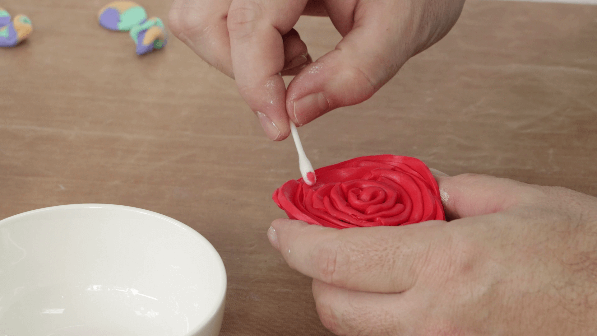 A red rose made from polymer clay, a hand uses a cotton bud tip to remove fingerprints from the clay.