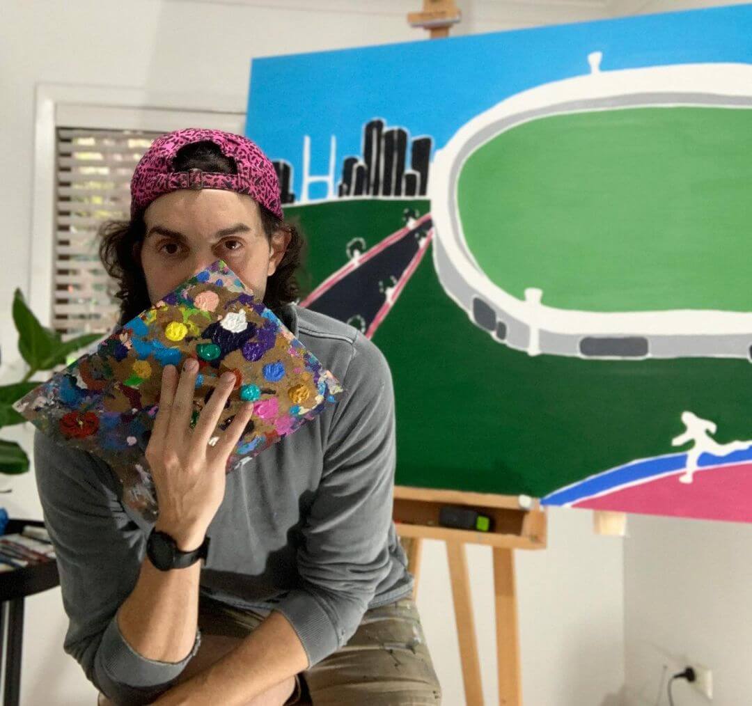 5. Artist Charles Nanopoulos holding a paint palette in front of his face with the MCG artwork behind him.