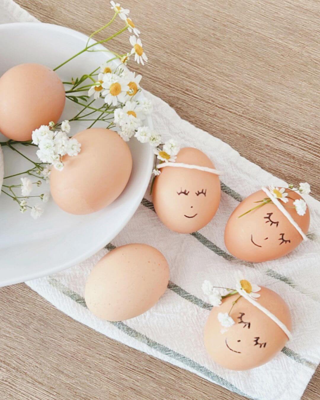 Eggs with cartoon faces drawn on them and small flower crowns. Eggs are on a towel on a wooden table.