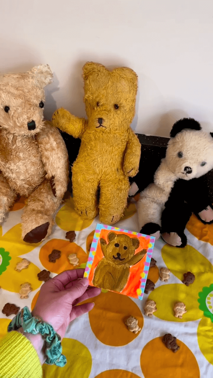 Hand holding a painting of a teddy bear next to three vintage teddy bears.