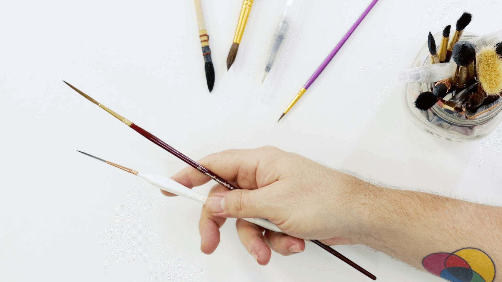 Hand holding two rigger watercolour brushes with several watercolour brushes around.