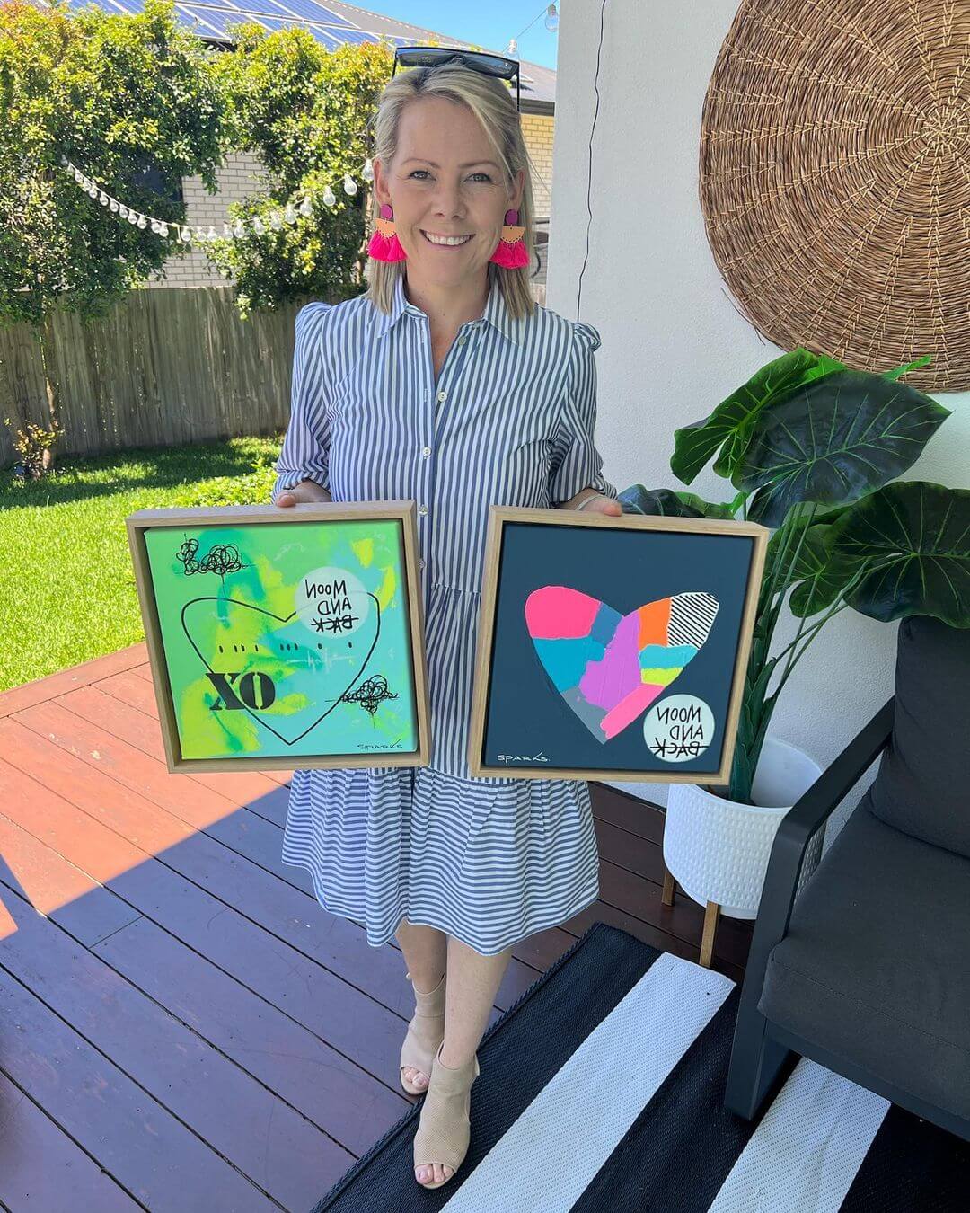 Michelle holding two of her artworks wearing a blue striped dress and pink earrings.