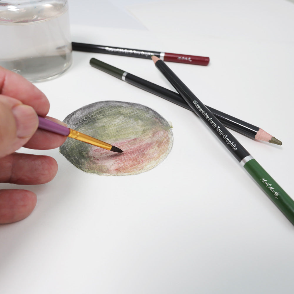 Three earth coloured watersoluble pencils lying next to paper as a watercolour brush activates the colours in a drawing.