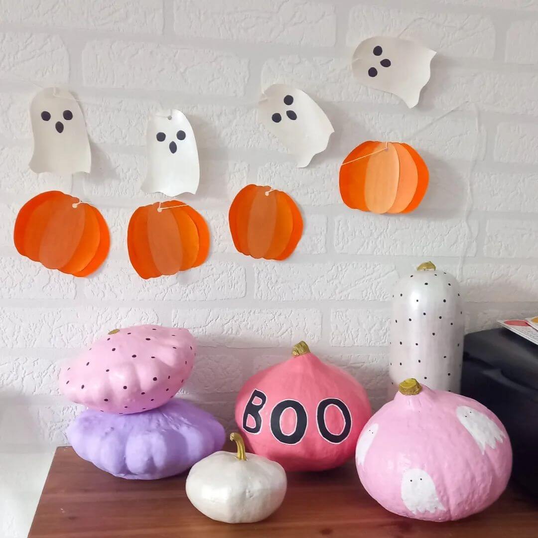 Various pastel painted pumpkins on a wooden table with pumpkin and ghost garland hanging above them.