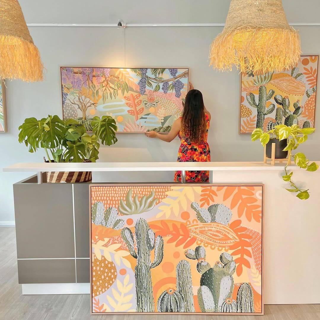 Zara hanging a nature landscape on a white wall with a cactus painting in front of her.