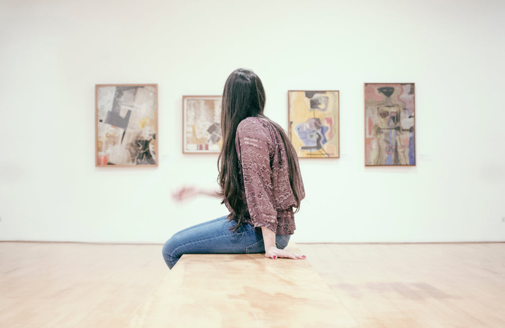 Woman sitting on wooden bench turning head to look at wall with 4 hanging abstract art works. 