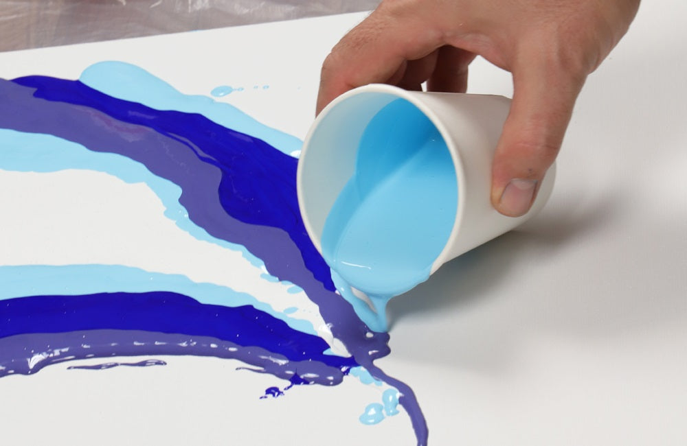 A plastic cup with light blue paint pouring on top of medium blue and purple, pour paints onto a canvas.