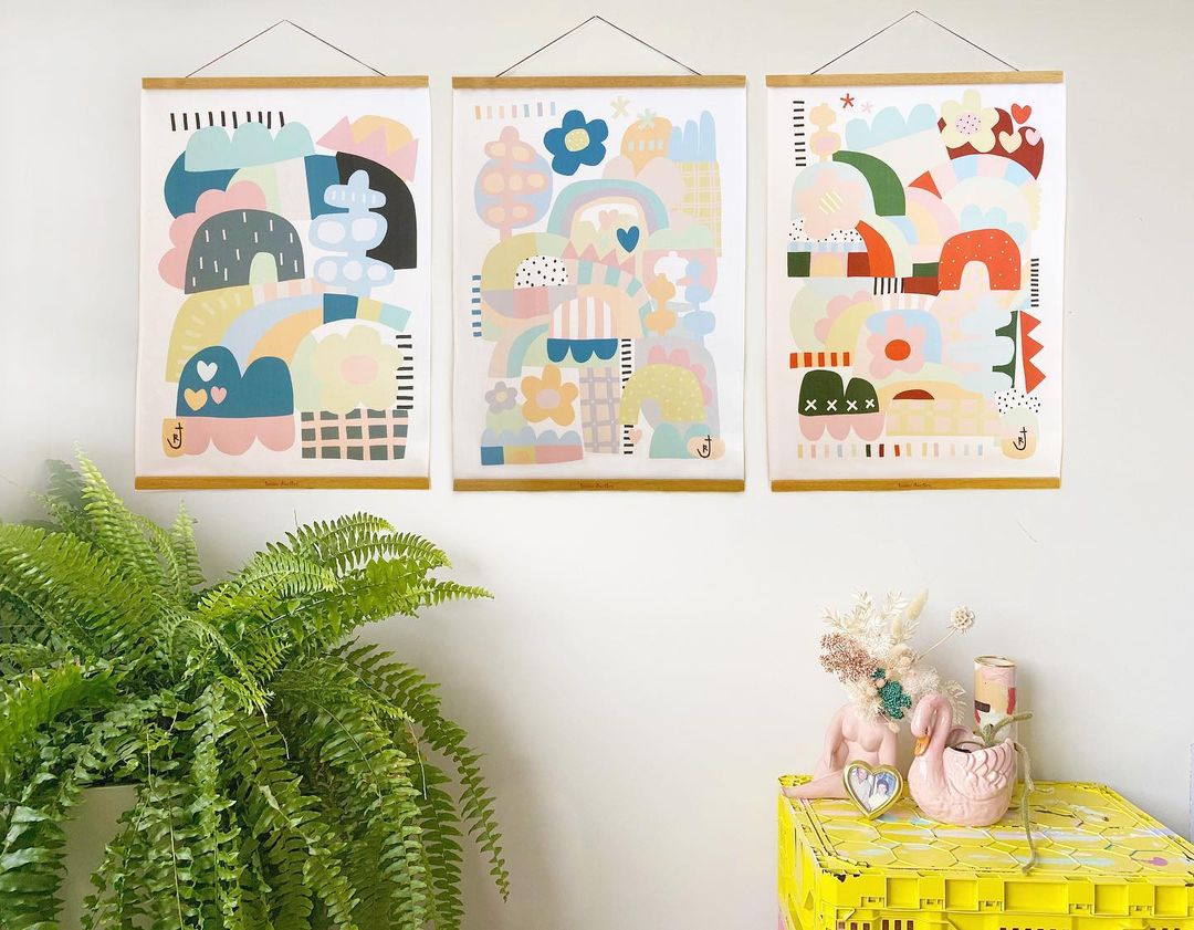 Three abstract wall hangings hanging on a wall near a yellow palette as a table and a plant.