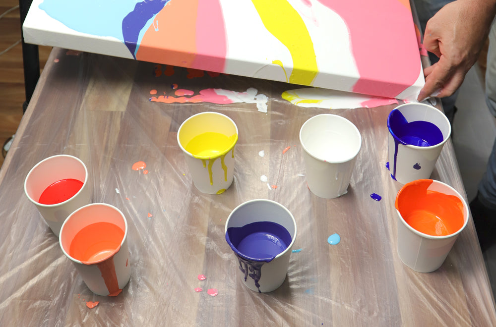 Cups filled with different coloured pouring paints, dripping over cup edges and onto plastic covered table.