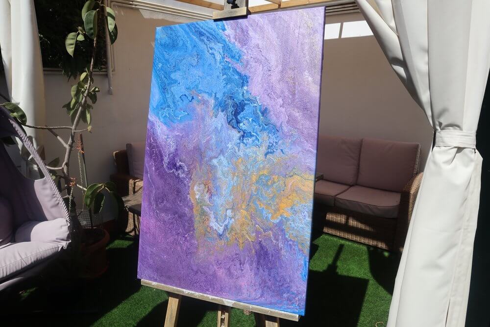 A blue, purple and gold pour painting on a wooden easel on a balcony outdoors.