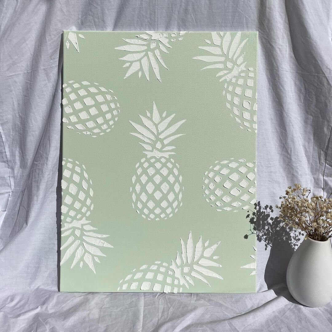 A green artwork with white pineapples printed on top, next to a vase with dried flowers.