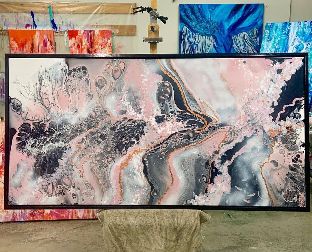 A large pour painting painted with black, pink, white, grey and gold paints mounted on an easel. The artwork has a black wooden frame around it with various colourful artworks.