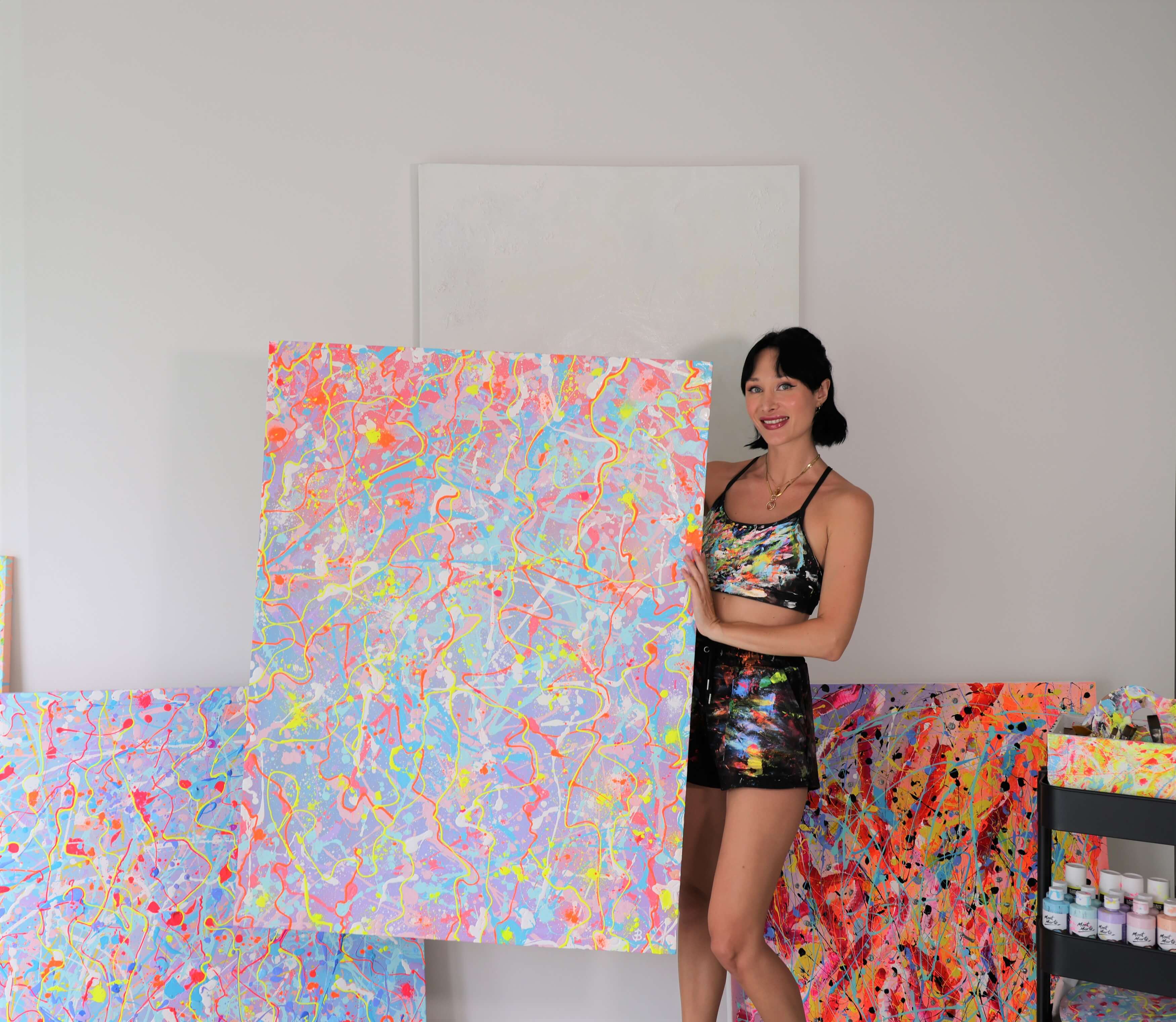 Artist Bridget holding a canvas smilling with abstract paintings behind her and an art trolley next to her