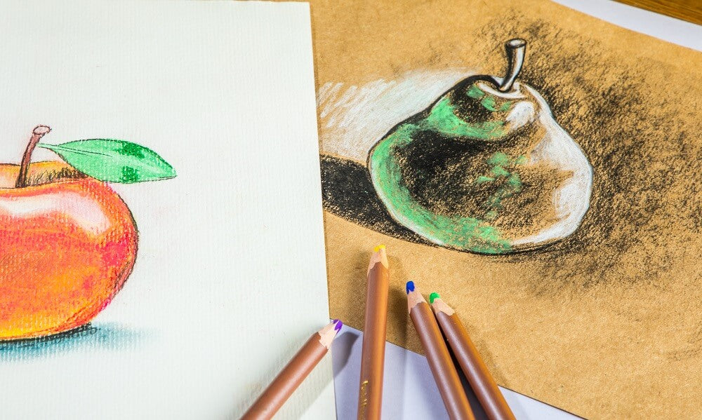 Four pastel pencils laying on top of a pastel artwork of a pear with a drawing of an apple next to it.