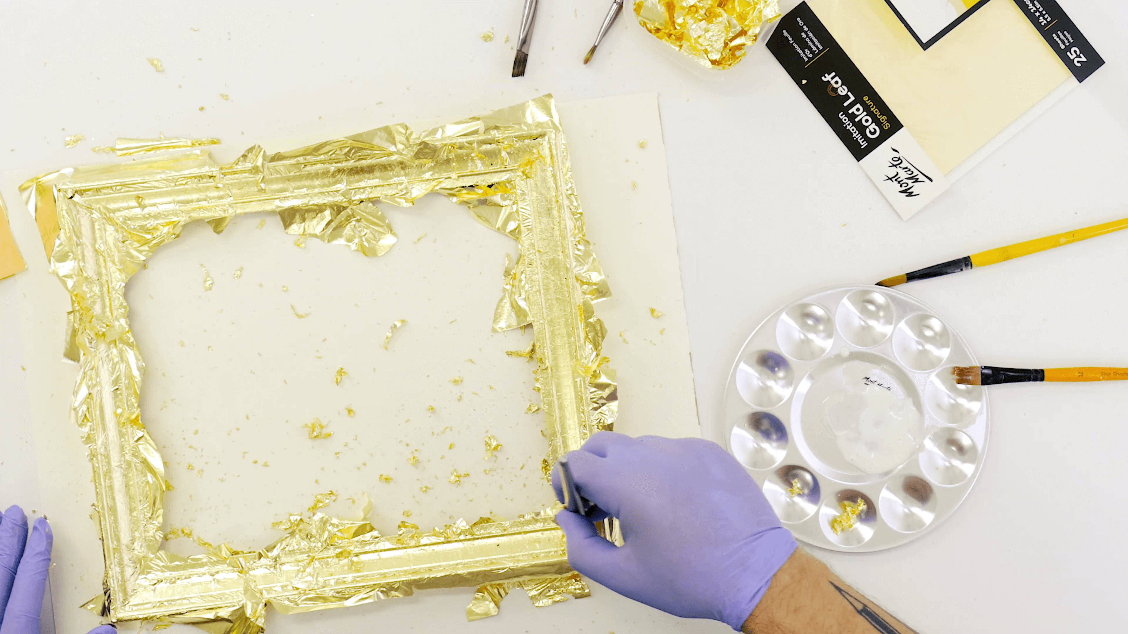 Purple gloves wrapping a wooden frame with gold leaf.