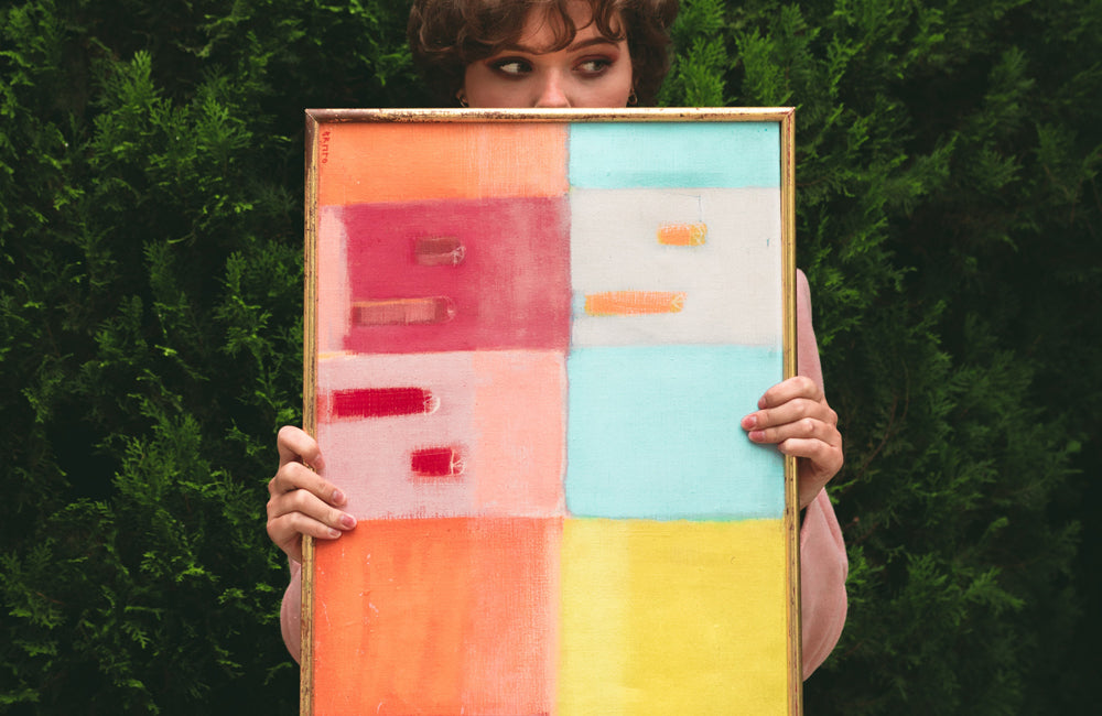 Woman holding pink, red, blue, orange and yellow coloured art work in front of body only showing her hands and top part of her face.  