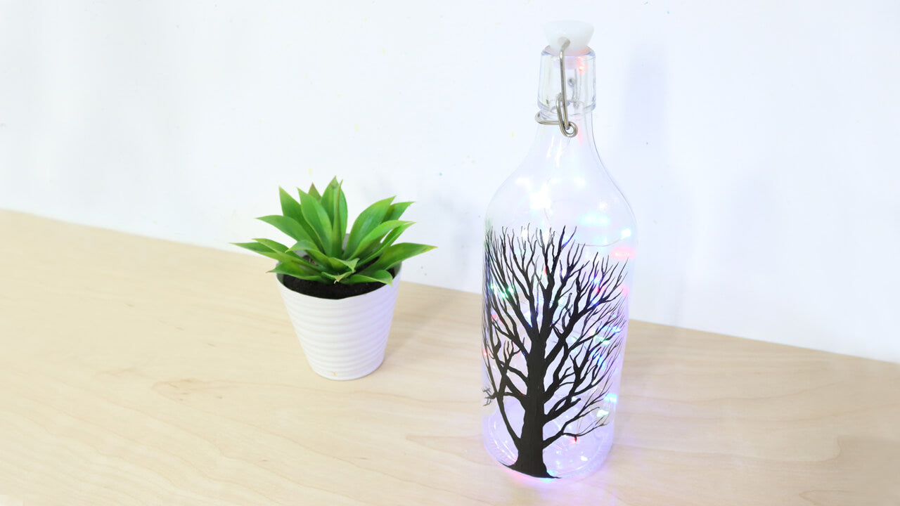 A recycled glass bottle turned into a lamp with tree silhouette and pot plant next to it.