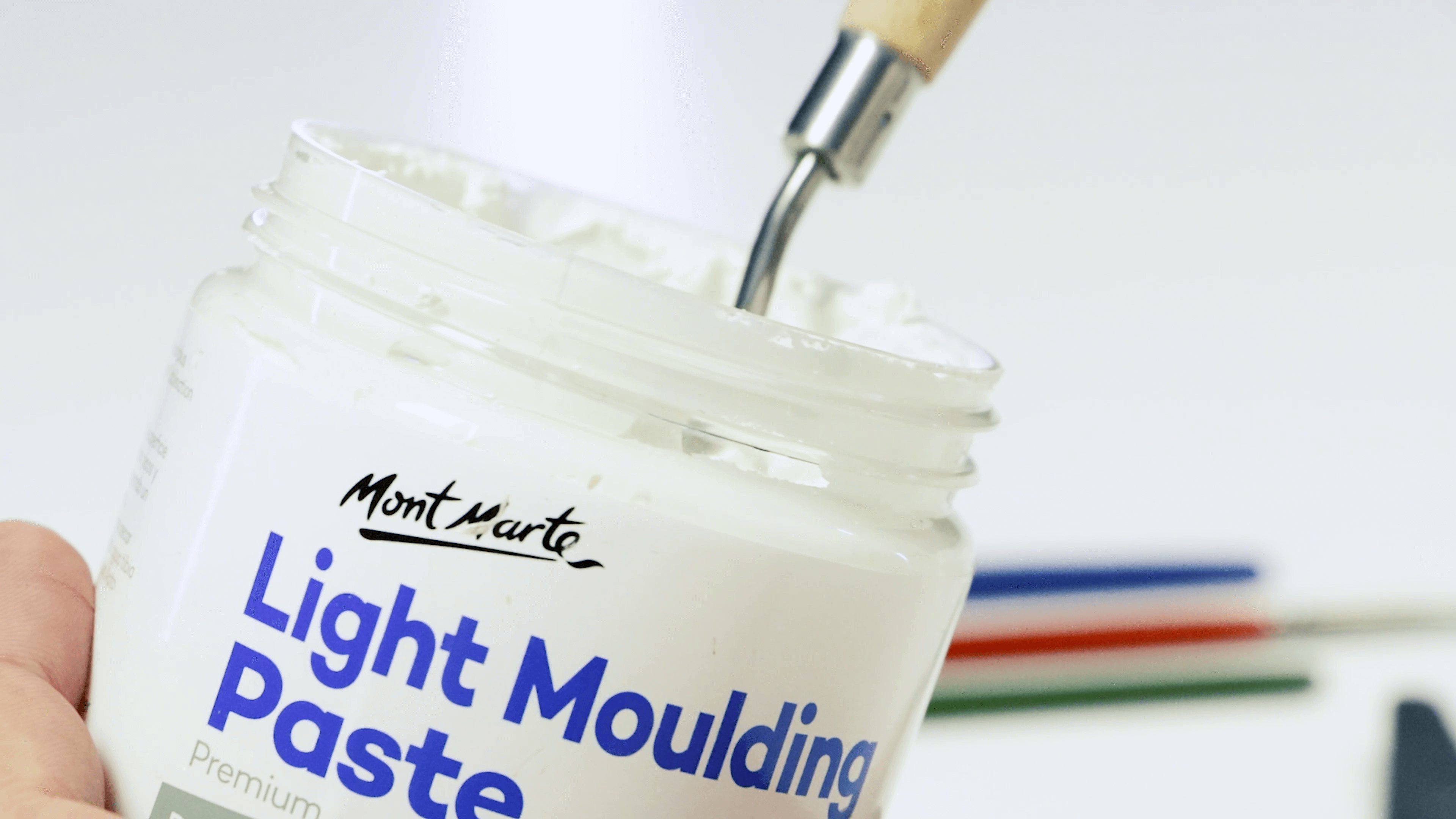 A tub of Mont Marte light moulding paste with a palette knife digging into the tub and a hand holding the tub on the left side.