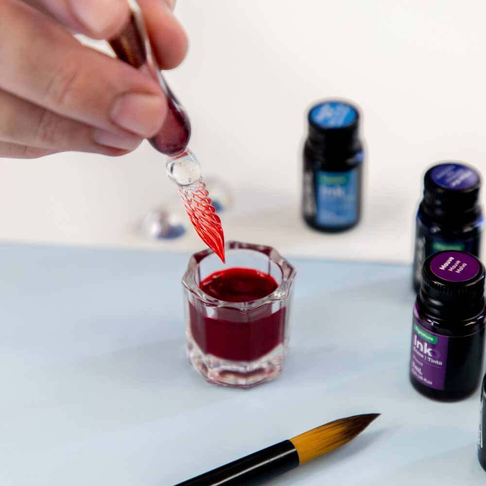 Hand holding a glass dipper pen about to submerge the pen into an ink well of red ink