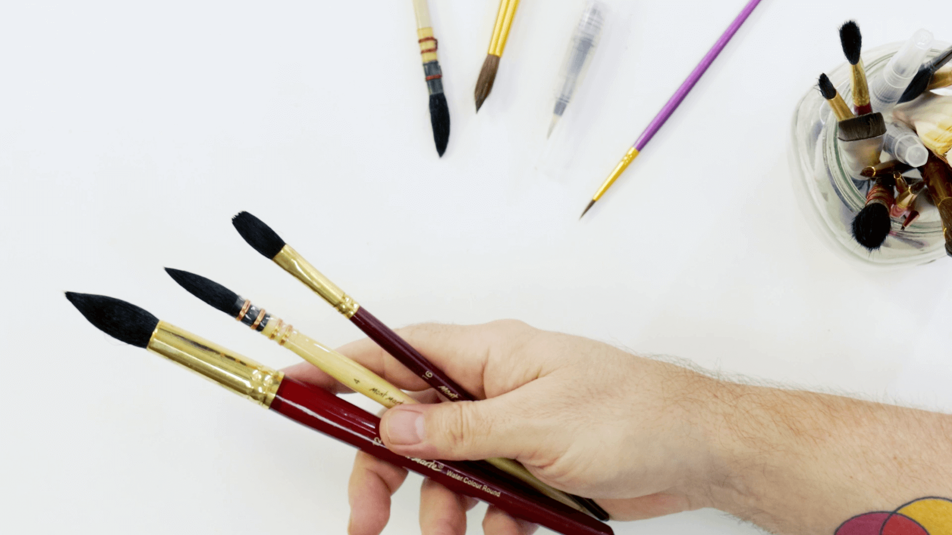 Hand holding three mop watercolour brushes with various watercolour brushes nearby.