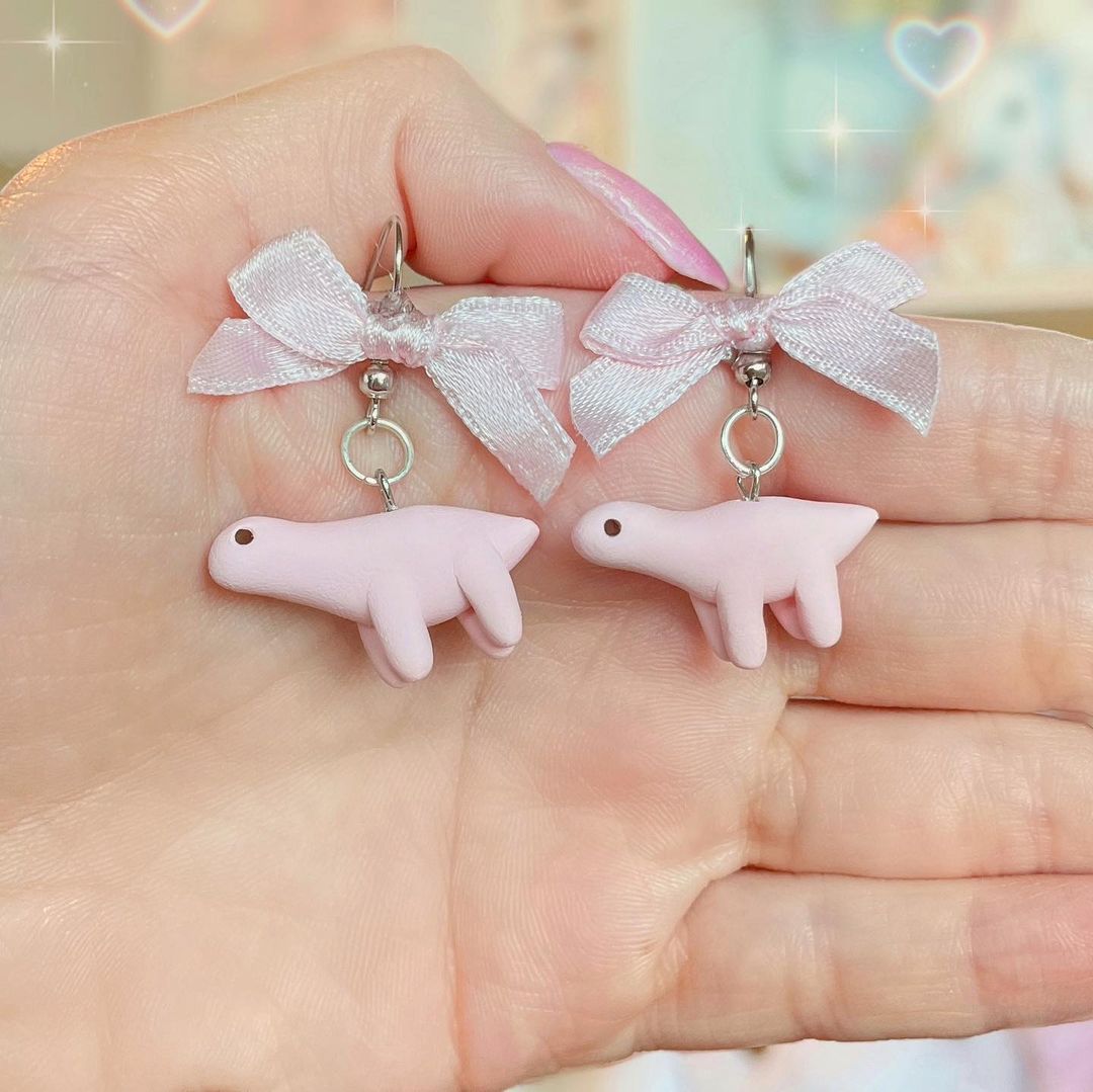 7. @olivescharms pink dinosaur scultpure earrings with bows on the stems, held on a hand with pink nails