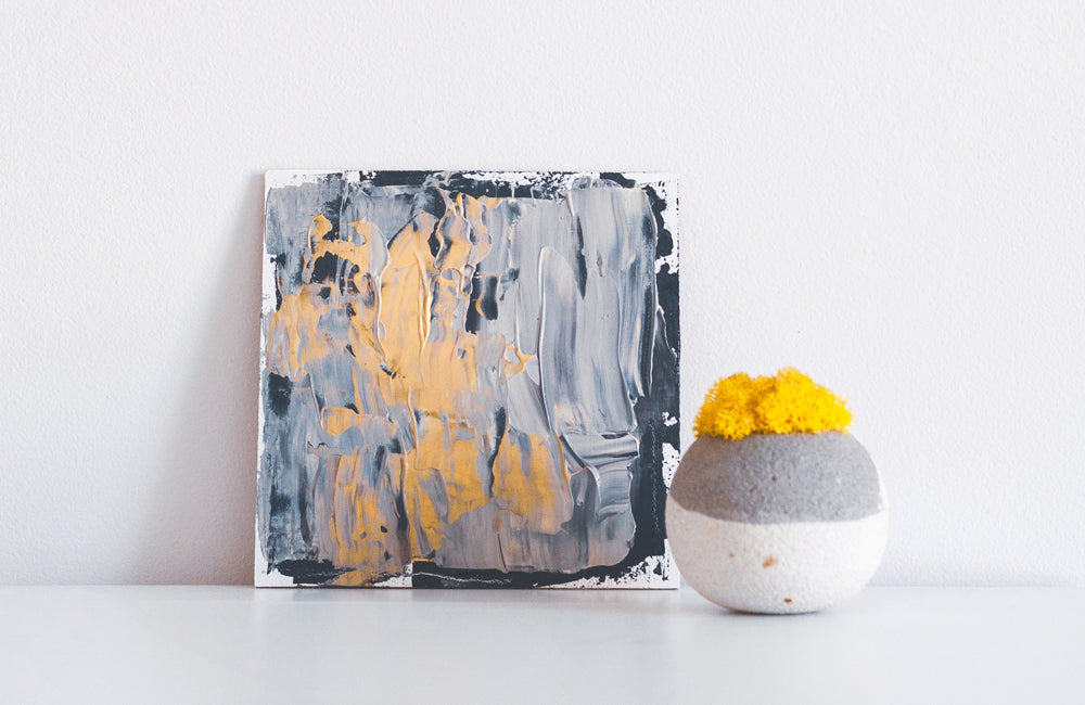 Pale orange, dark and light grey painting on square canvas next to a grey and white sphere vase of yellow marigold flowers. 